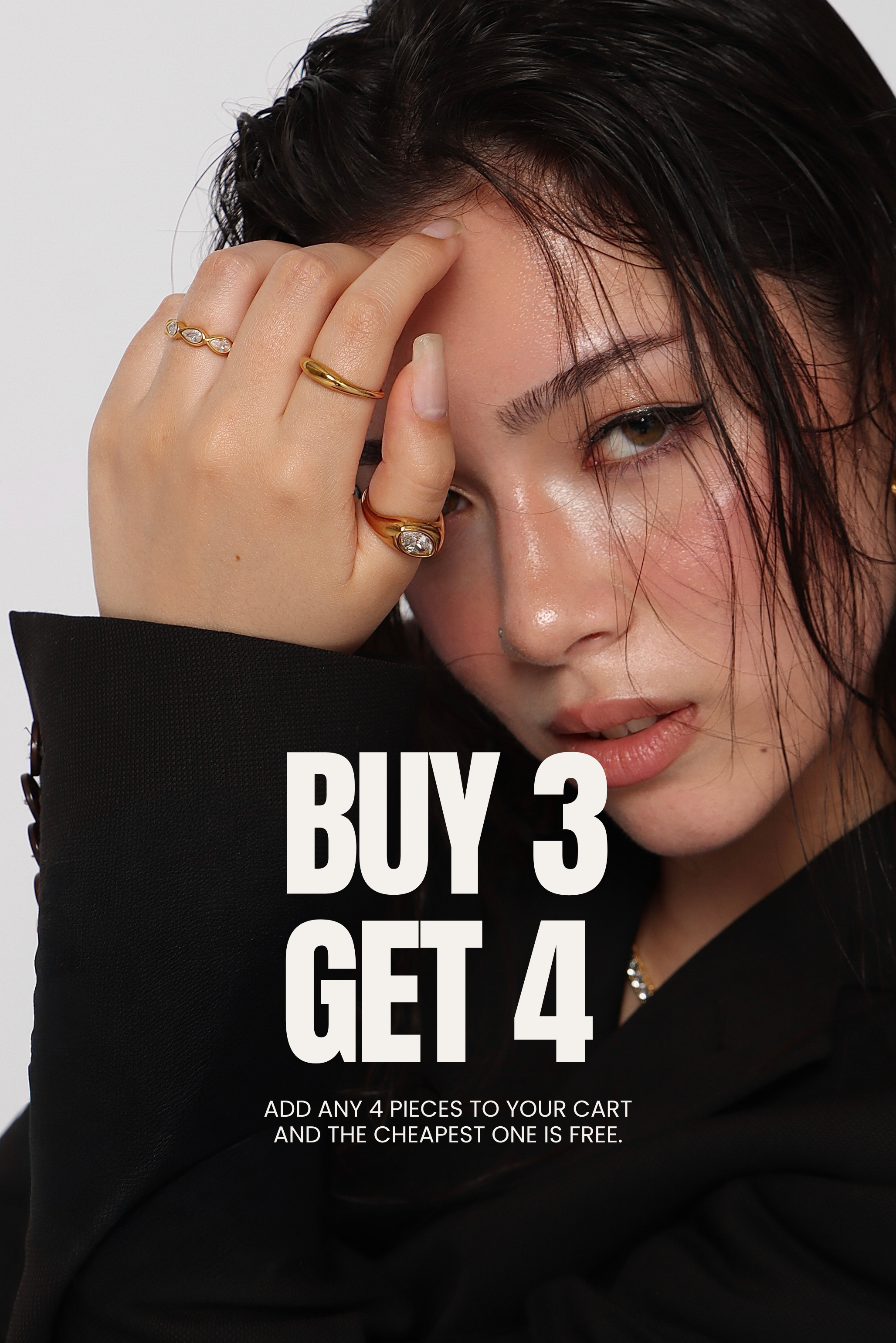 A close-up promotional photo of a woman with dark hair and makeup, wearing rings, with large white text reading 'BUY 3 GET 4' and smaller text below over a black garment against a light gray background.