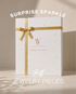 A white rectangular gift box with a gold ribbon and bow, decorated with small gold star accents, standing on a beige surface. White text above reads “SURPRISE SPARKLE” and below reads “24 JEWELRY PIECES.